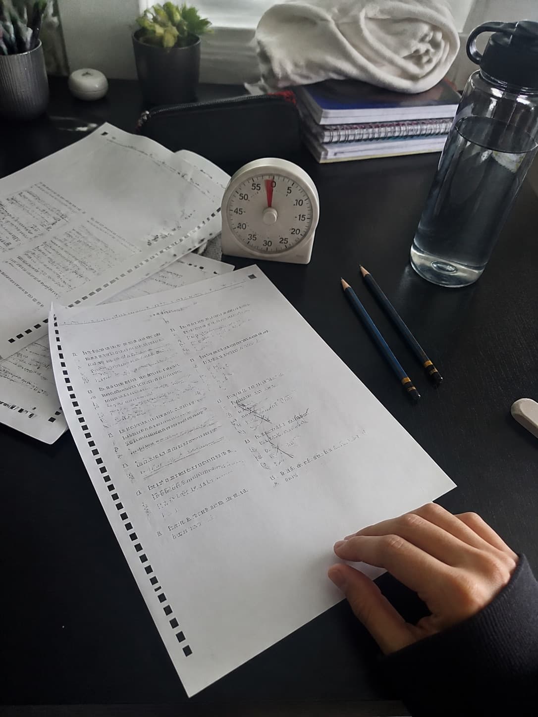 A desk after a timed mock, with used papers, an analogue timer, pencils and a water bottle
